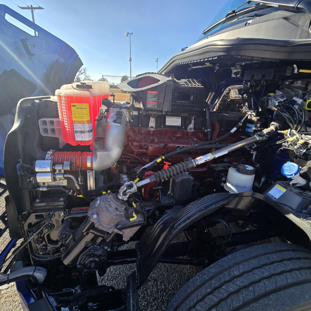 Engine bay of a 2026 Mack PR64T76H truck showing coolant reservoir with red cap, various hoses, wiring, and turbo area.