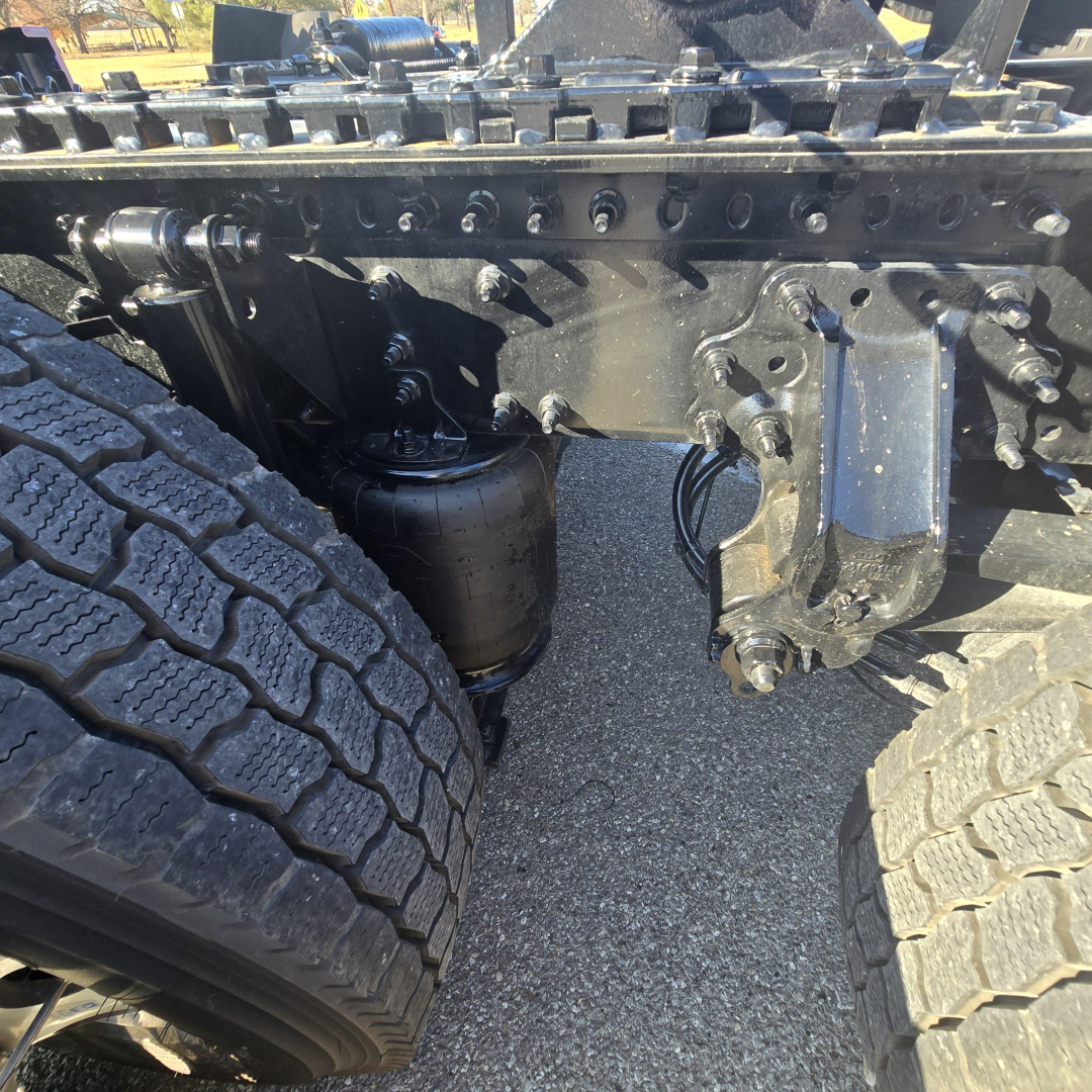 Mack PR64T76H truck front suspension showing dual rear drive tires, leaf springs, and air suspension canister with surrounding frame components visible.