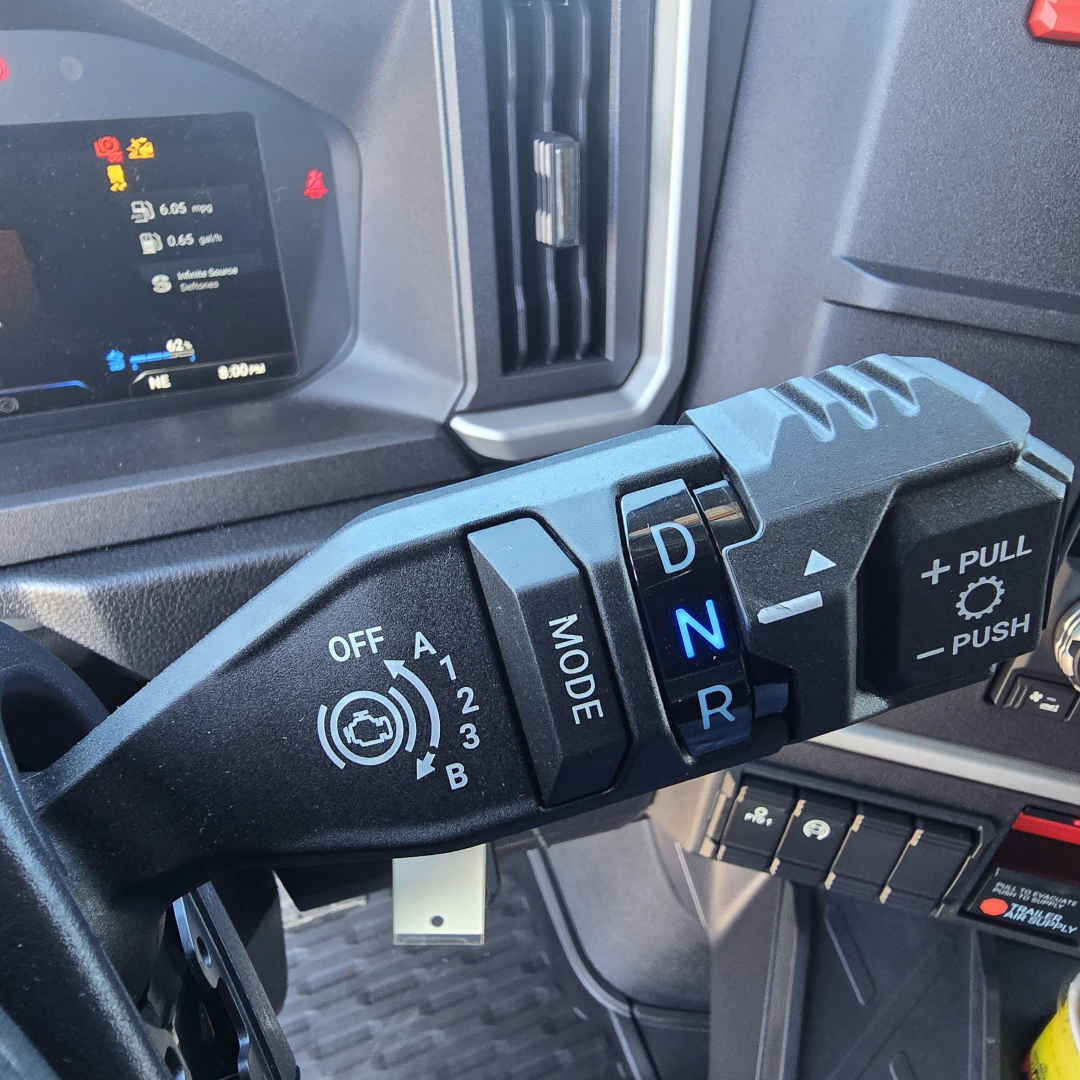 Cab interior of a 2026 Mack PR64T76H showing gear selector stalk with D N R labels and mode switch, dashboard display in background.