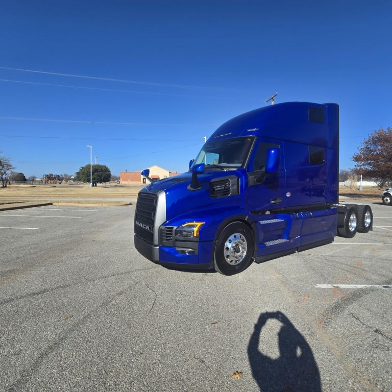 Truck cab close-up showing the blue Mack PR64T76H sleeper tractor with front grille, headlights, and side mirrors in clear view.