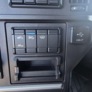 Dashboard controls panel with four labeled switches and icon indicators, plus a small USB-C port cover to the right; navigation slot below.