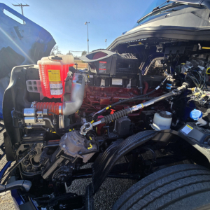 Engine bay of a 2026 Mack PR64T76H truck showing coolant reservoir with red cap, various hoses, wiring, and turbo area.
