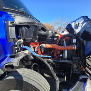 Engine bay of a blue Mack PR64T76H truck showing orange coolant hose, blue fuel lines, radiator and engine components.