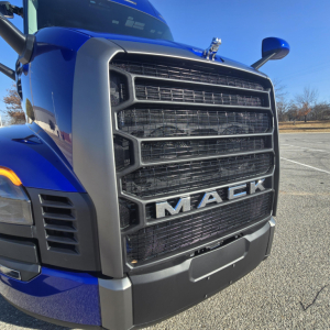 Truck grille front view of a blue 2026 Mack PR64T76H showing large black radiator grille with MACK logo and chrome accents.