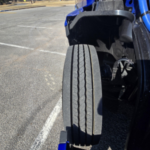 Front left dual-tire setup of a blue Mack PR64T76H truck showing a large steer axle tire with tread pattern and surrounding components