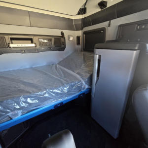 Interior sleeper cab of a 2026 Mack PR64T76H truck showing a bunk bed with plastic-covered mattress, side cabinet, and dashboard controls.