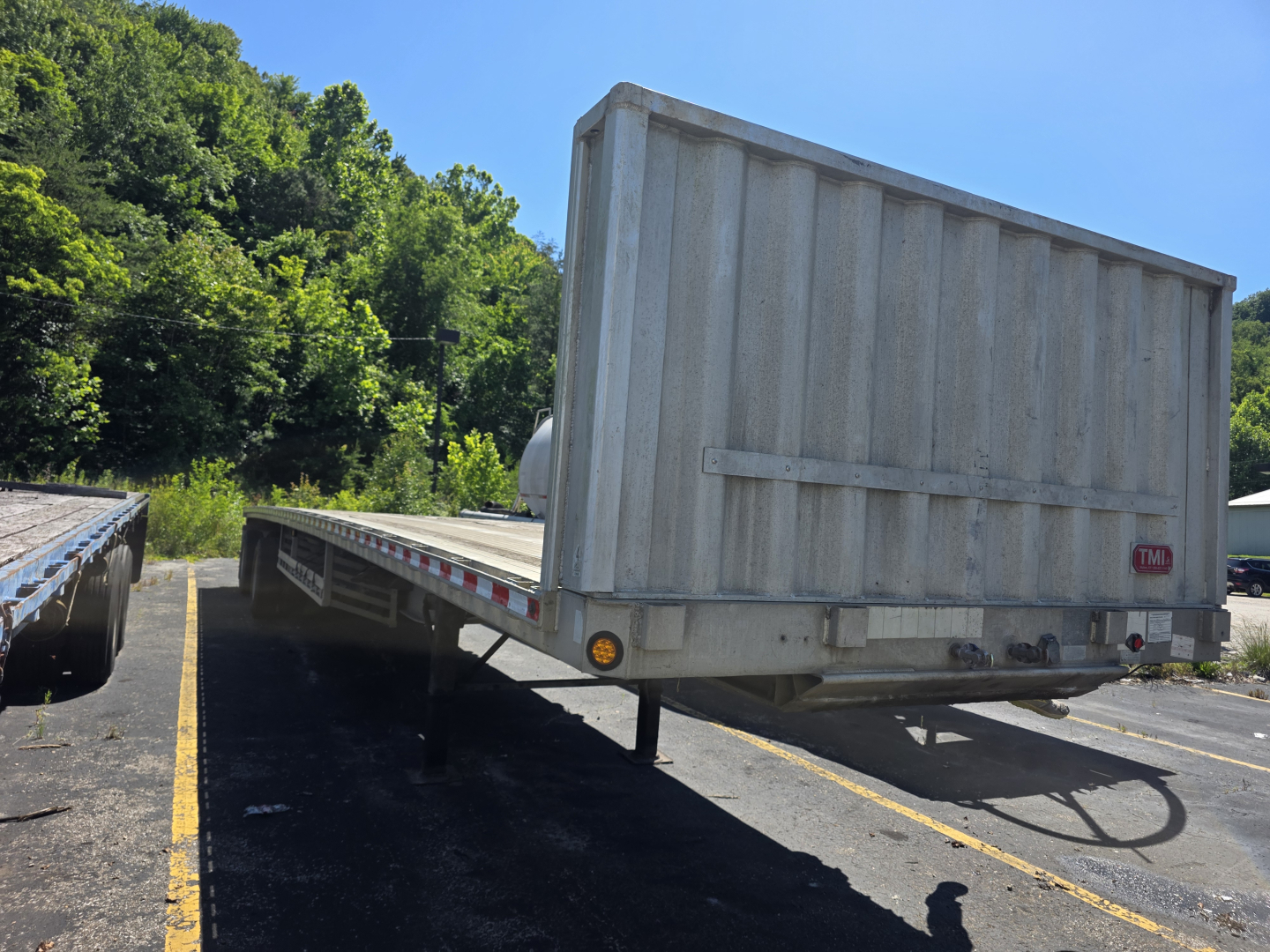 Flatbed trailer with metal ramp and rear access, Fontaine Phantom trailer in a parking lot, daylight, green trees in background.