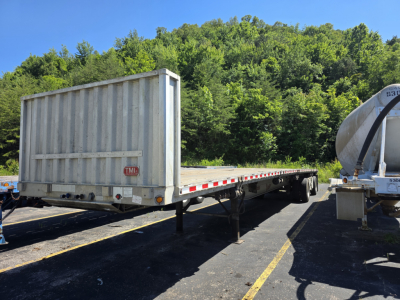 Plain rectangular flatbed trailer with raised end gates, used Fontaine Phantom trailer in a parking lot with trees in background.