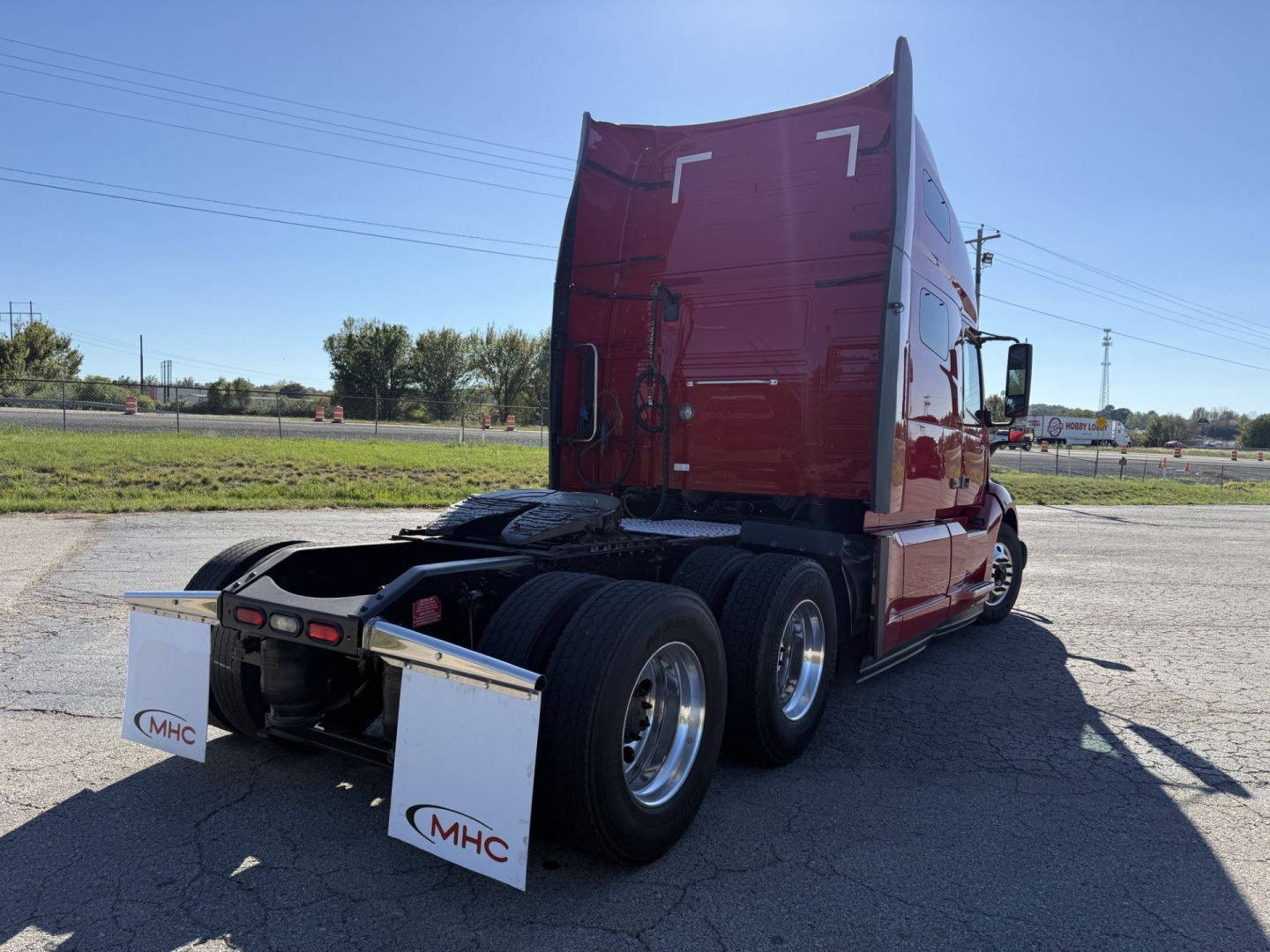 Rear view of a red Volvo VNL64T760 semi-truck tractor with sleeper cab, showcasing rear axles and mud flaps with MHC logos.