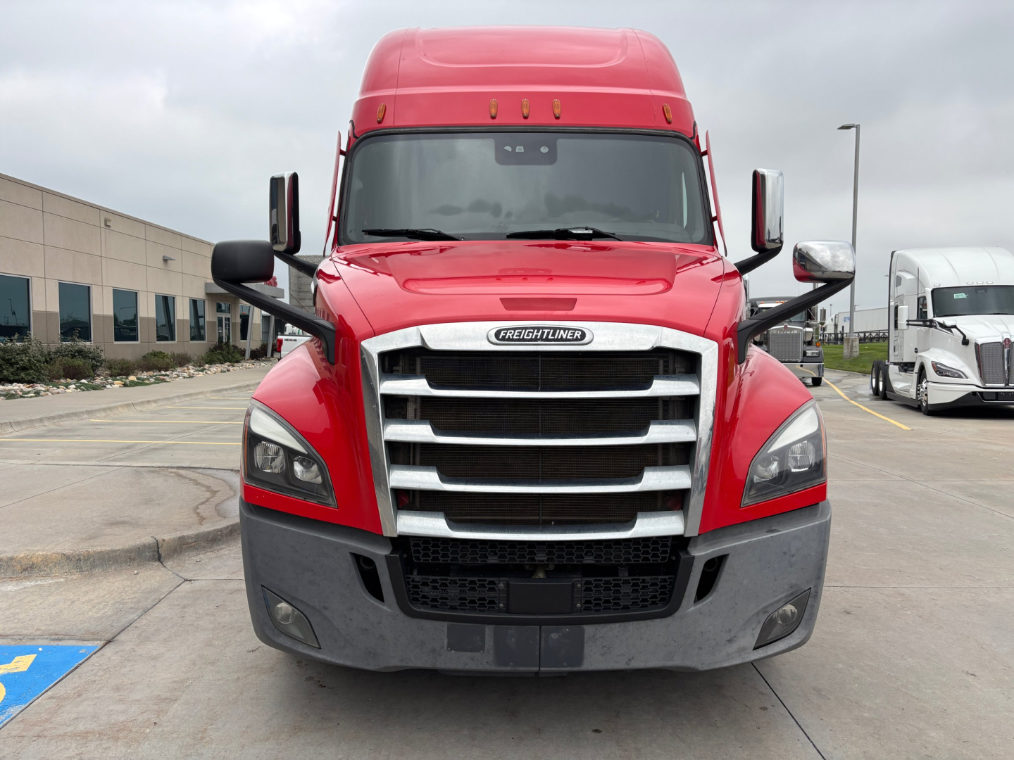 Truck front view showing Freightliner emblem, large chrome grille, red cab, black bumper and side mirrors.