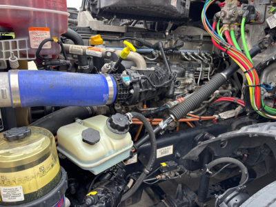 Truck engine bay with multiple colored hoses, wiring, reservoirs, and a blue intercooler hose. Visible fluid reservoirs and engine components in a Freightliner PT126064ST truck.
