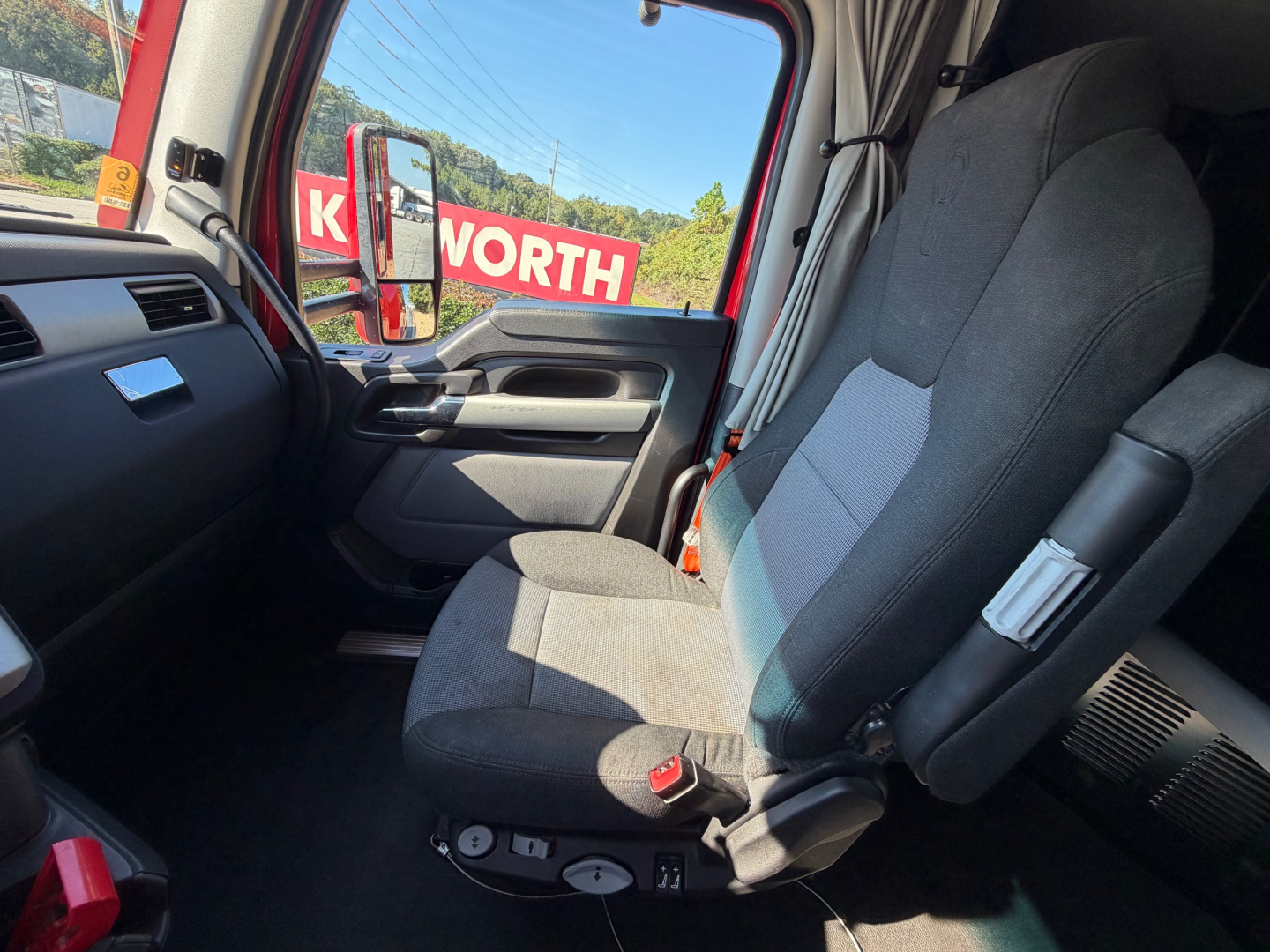Kenworth T680 cab interior showing driver seat and dashboard with window view; partial Kenworth dealership sign visible through window.