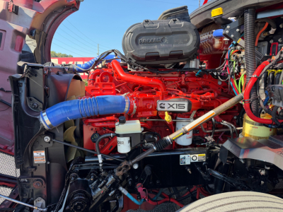 Kenworth T680 engine bay with bright red engine, blue intercooler hose, black plastic air intake cover, white reservoirs, and various hoses and wiring visible.
