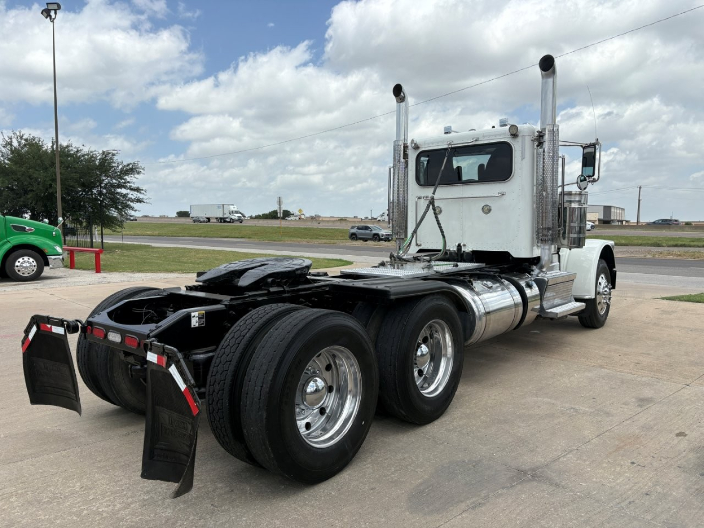 Peterbilt 389 daycab tractor with dual rear axles and chrome fuel tanks, front cab, exhaust stacks, and mud flaps visible in a parking lot.
