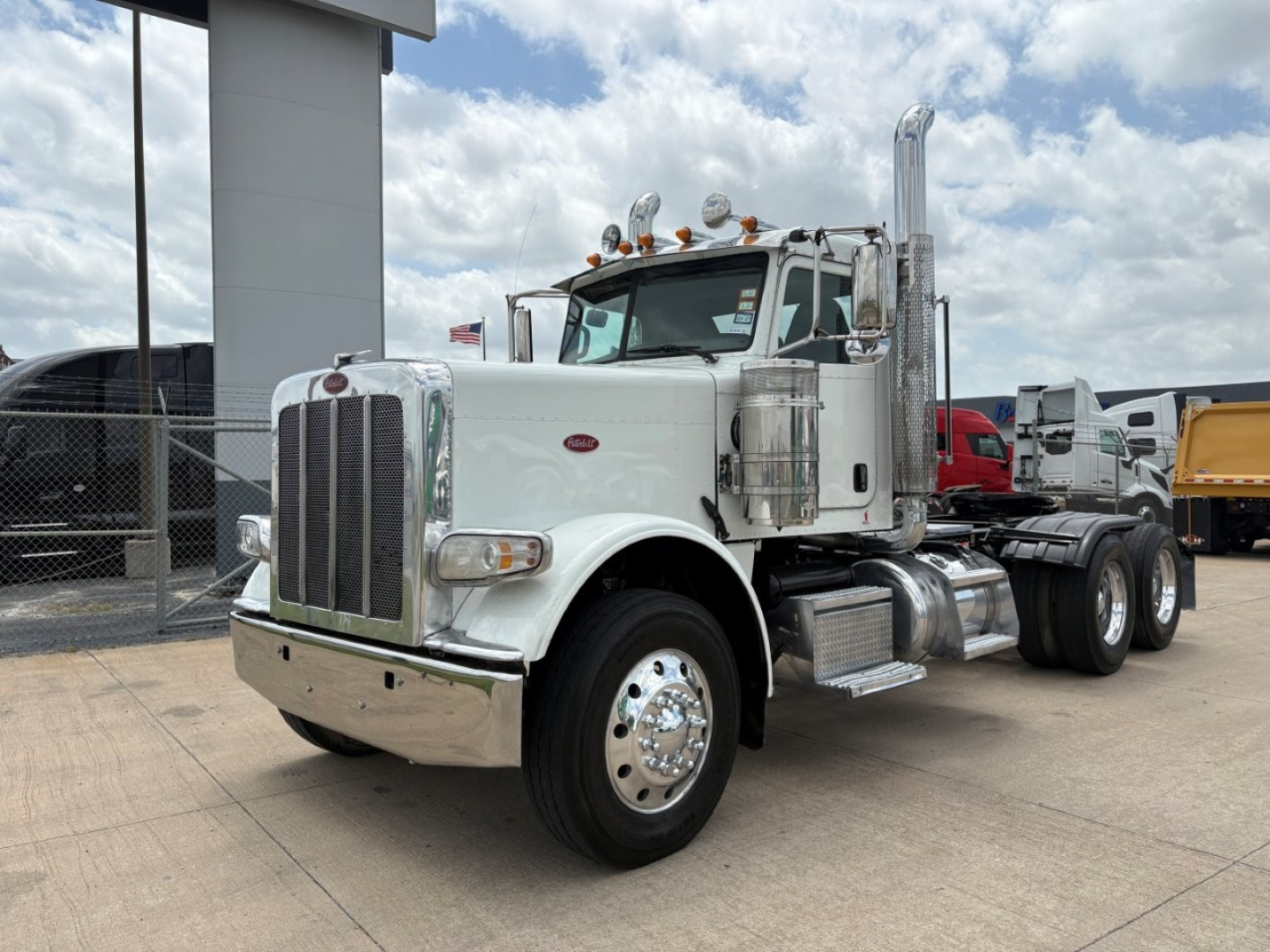Peterbilt 389 conventional daycab truck in a dealership lot, front three-quarter view showing chrome grille, dual exhaust stacks, mirrors, and tandem rear axles.
