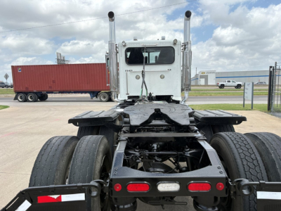 Peterbilt 389 conventional daycab chassis rear view showing dual rear axles and fifth wheel mounting, white cab, with white and red taillights, trailer in background.