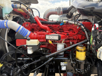 Peterbilt 389 engine bay showing 6-cylinder red diesel engine with X15 badge and various hoses and reservoirs.