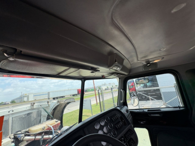 Peterbilt 389 truck interior cab view showing instrument cluster, dashboard, and overhead storage. Exterior scene through windshield with chain-link fence and equipment.