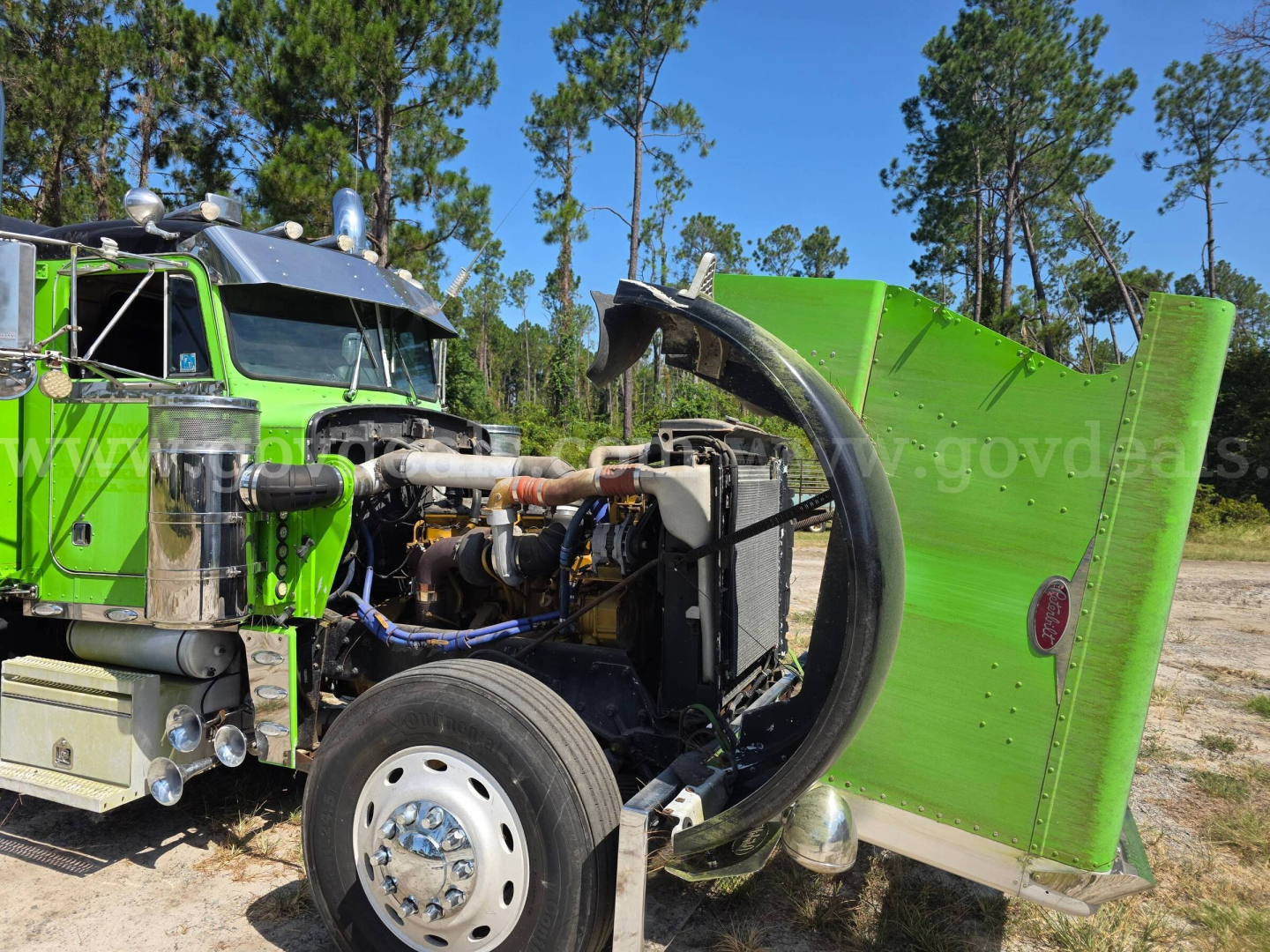 Peterbilt 359 truck with bright green cab and exposed engine bay; large curved exhaust pipe and engine components visible; front wheel and fender; open hood/hood support exposing mechanical parts.