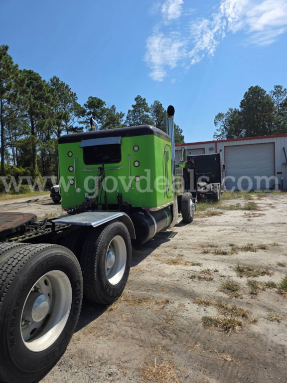 Green Peterbilt truck cab on trailer chassis in an outdoor lot, with a blue sky and trees in background.