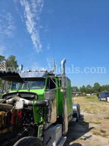 Truck front-left view of a green 1986 Peterbilt 359 with exposed engine, chrome exhaust, and cab in a yard.