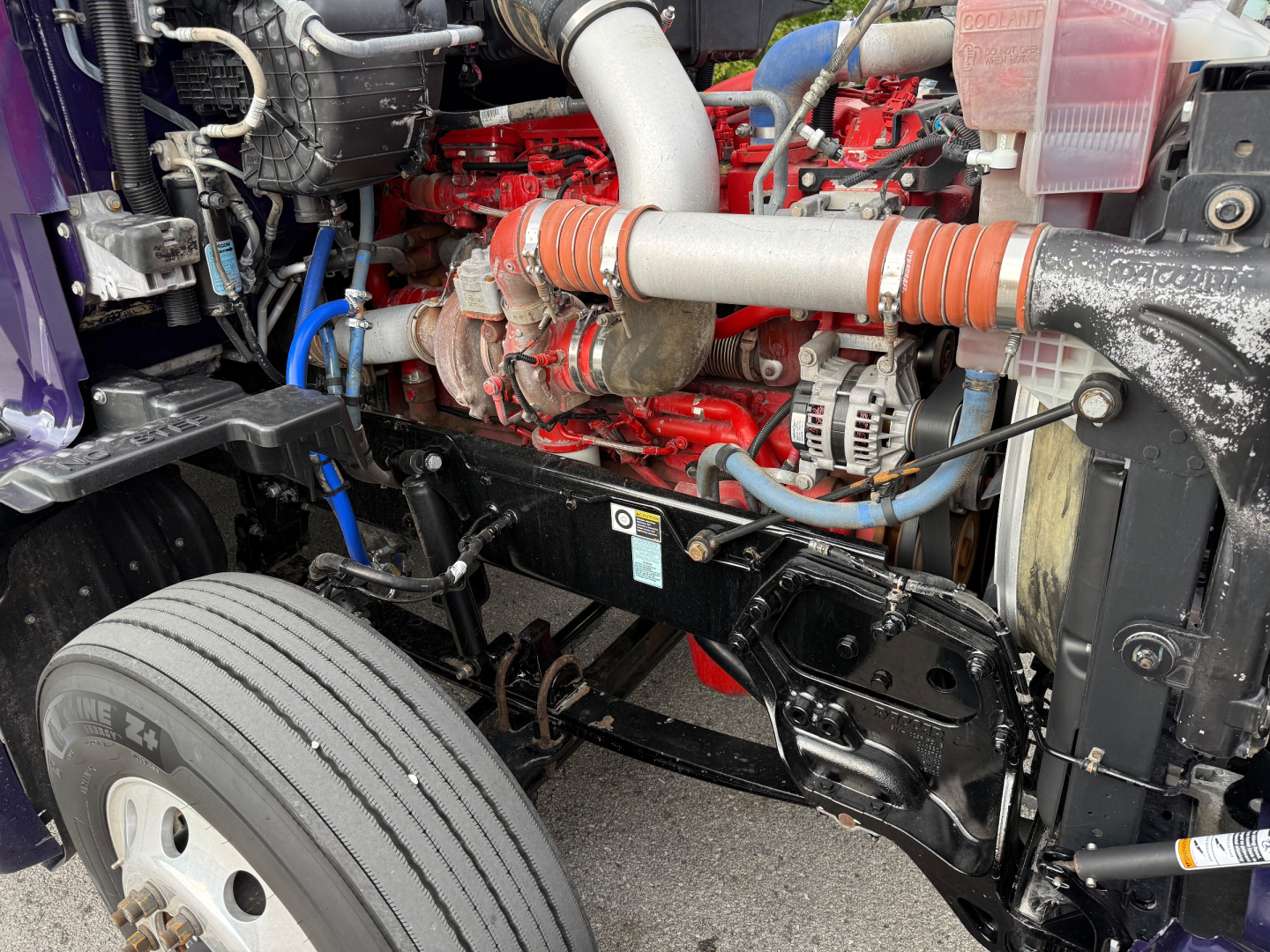 Kenworth T680 engine bay showing red engine block, hoses, coolant reservoir, and front suspension components.