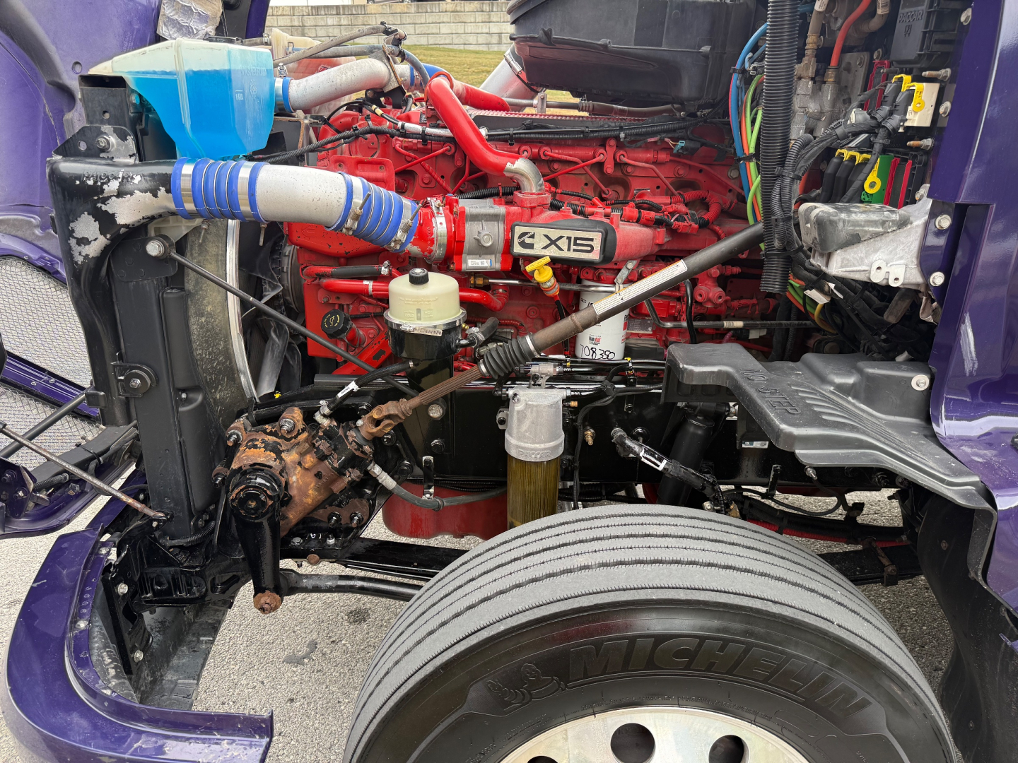 Kenworth T680 engine bay showing red Cummins X15 engine with blue radiator hose, purple cab, and exposed components.