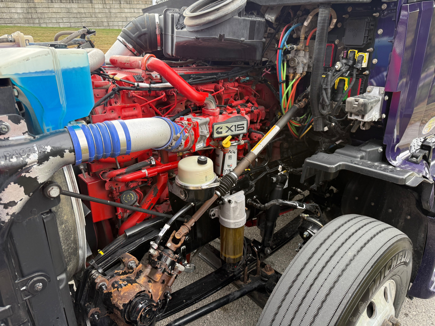 Kenworth T680 engine bay showing red engine with X15 badge, blue coolant reservoir, hoses, wiring, and front wheel.