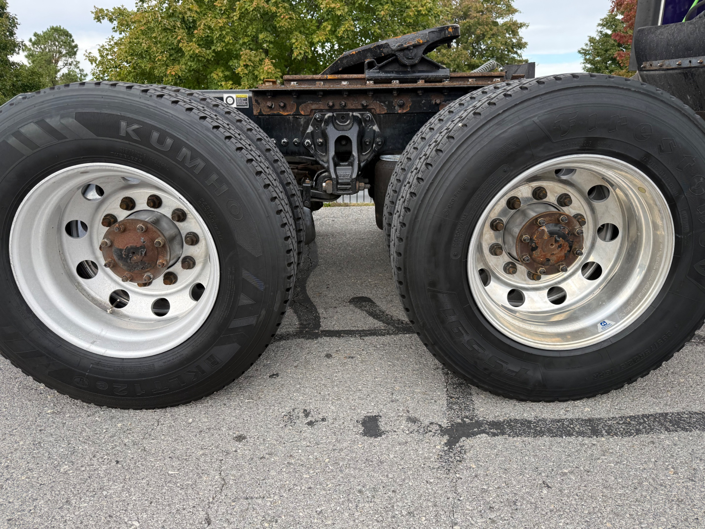 Dual rear tractor wheels and axle of a Kenworth truck showing aluminum rims and tire treads, with frame and fifth wheel mount visible between wheels.