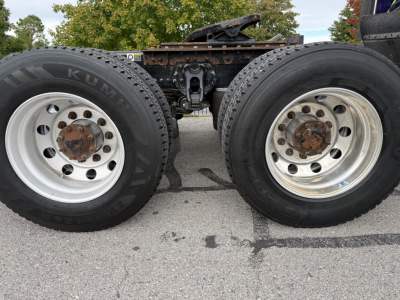 Dual rear tractor wheels and axle of a Kenworth truck showing aluminum rims and tire treads, with frame and fifth wheel mount visible between wheels.