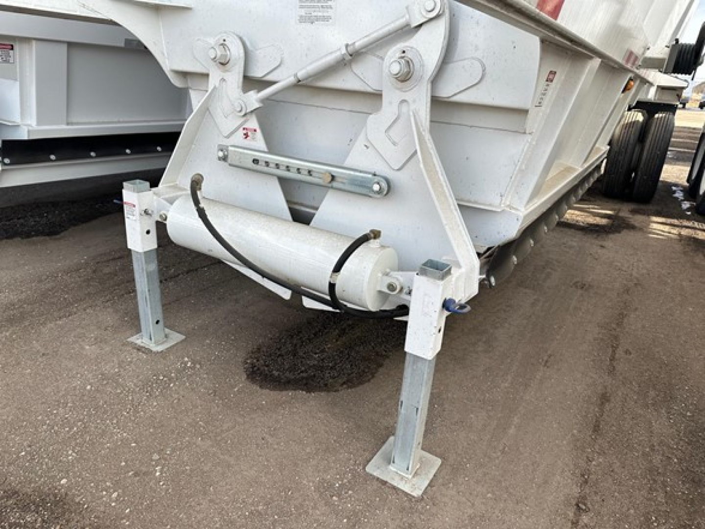 Bottom dump trailer tongue area with support legs and hydraulic cylinder visible, metal frame and ground beneath.