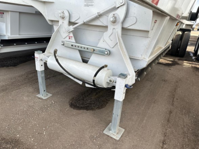 Bottom dump trailer hitch area showing hydraulic cylinder and landing legs with square bases in front; white-painted metal frame and undercarriage with gravel surface.