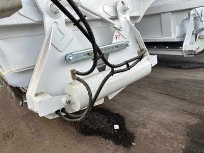 Bottom-dump trailer hitch area with hydraulic lines and support bars, white paint, dark spilled fluid on ground near frame.