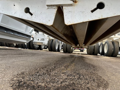 Bottom-dump trailer underside view showing hxed metal frame and tailgate with keyhole cutouts; gravel and tires in background.