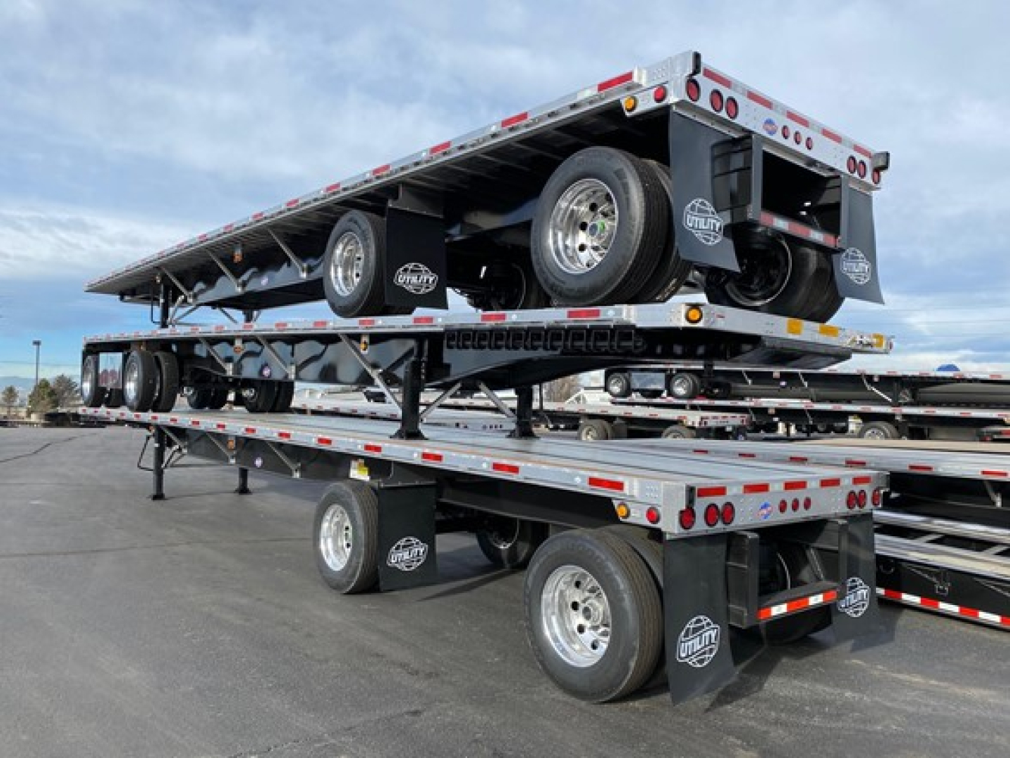 Flatbed trailer stacked two high showing two axles, spare tire, and reflective safety stripes in a trailer lot.