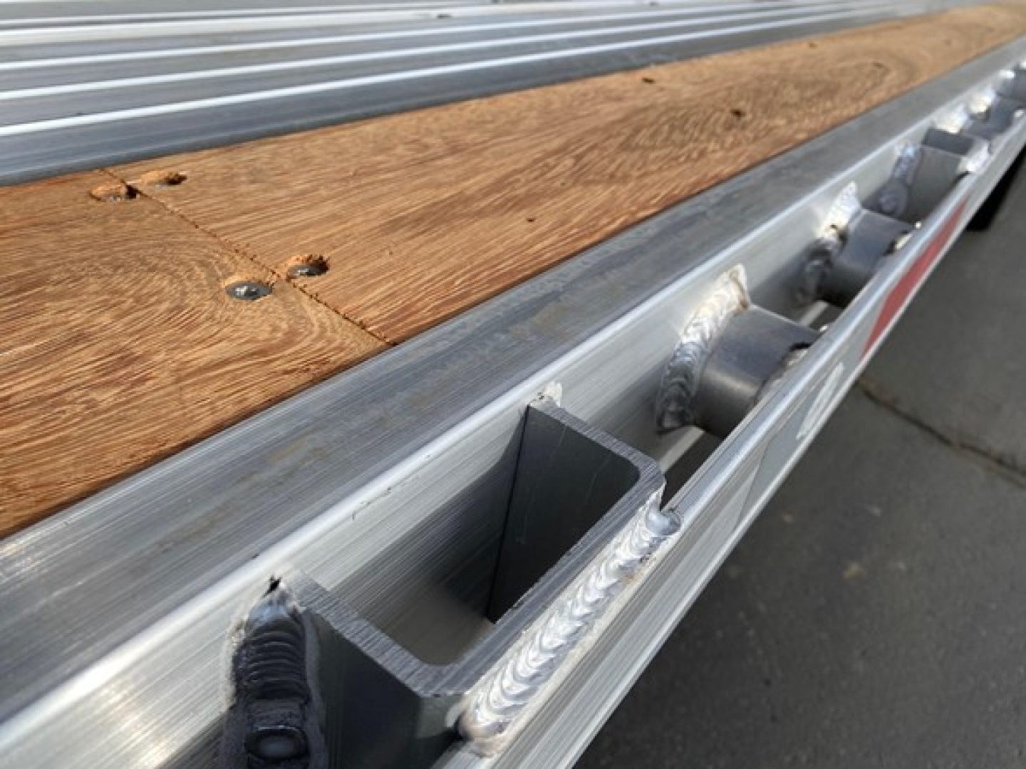 Trailer bed edge with wooden plank and side rail, view of aluminum frame and riveted brackets along the edge.