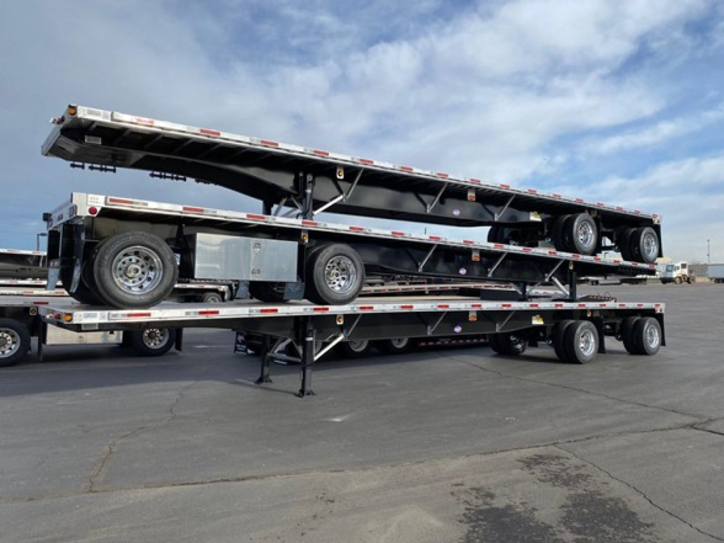 Flatbed trailer loaded on dual-level carrier with a folded gooseneck. Visible text on side rails includes safety stripes and reflective tape; no legible branding or model text discernible.