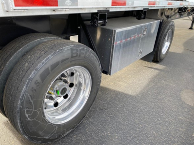 Trailer wheel and storage box under flatbed showing BFGoodrich tire and aluminum toolbox with reflective strip on rear.