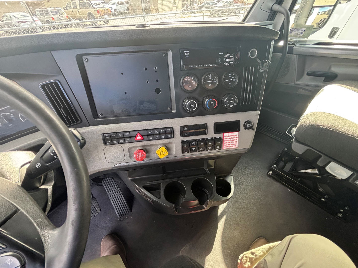 Cab interior of a Freightliner truck showing dash with gauge cluster, control switches, and steering wheel readable; cup holders and center console visible.