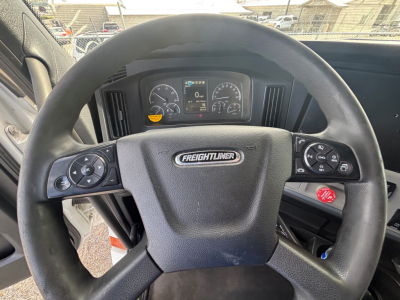 Truck steering wheel with Freightliner logo in open cab of a 2021 Freightliner CA-126 showing instrument cluster and control buttons on steering wheel.
