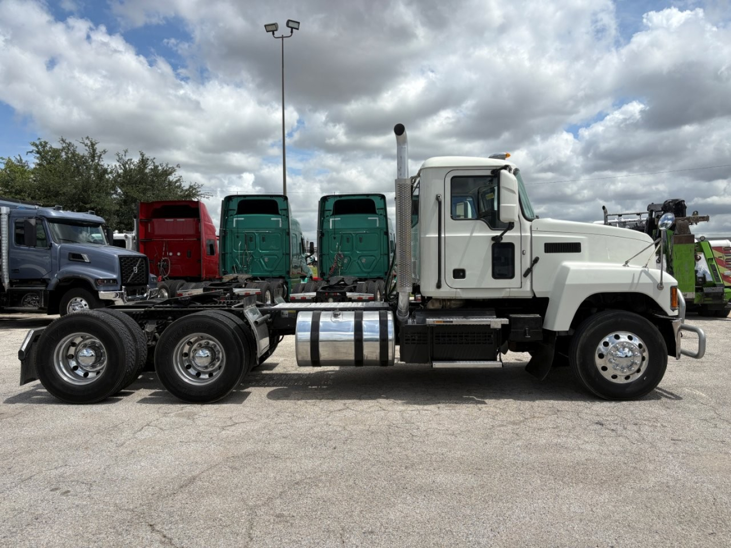 Truck chassis with white Mack PI64T cab, fuel tanks, and dual rear axles; other trucks in background.