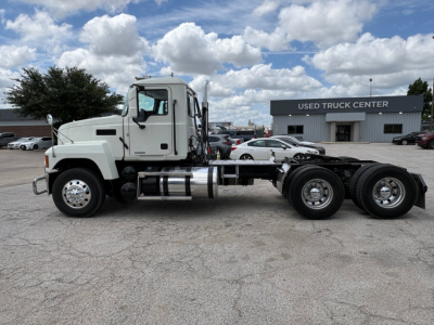 Side view of a white Mack PI64T truck chassis with cab and dual rear axles, fuel tanks, and exhaust; in background a building reading 'USED TRUCK CENTER'