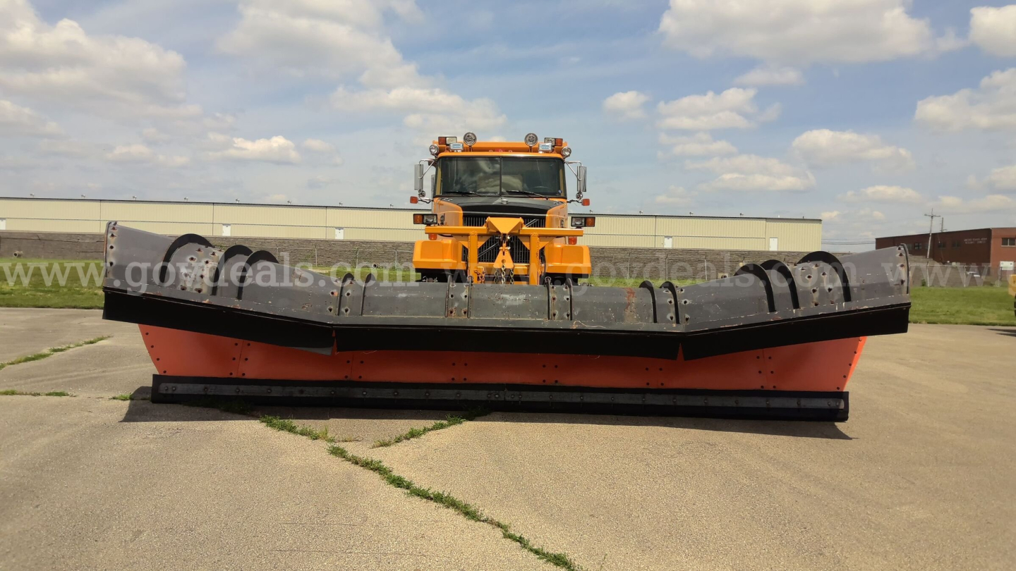 Volvo Autocar plow truck with large orange front-mounted plow and heavy-duty blade, wide tires, warehouse backdrop, clear sky.