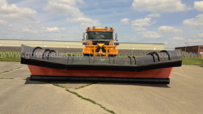 Volvo Autocar plow truck with large orange front-mounted plow and heavy-duty blade, wide tires, warehouse backdrop, clear sky.