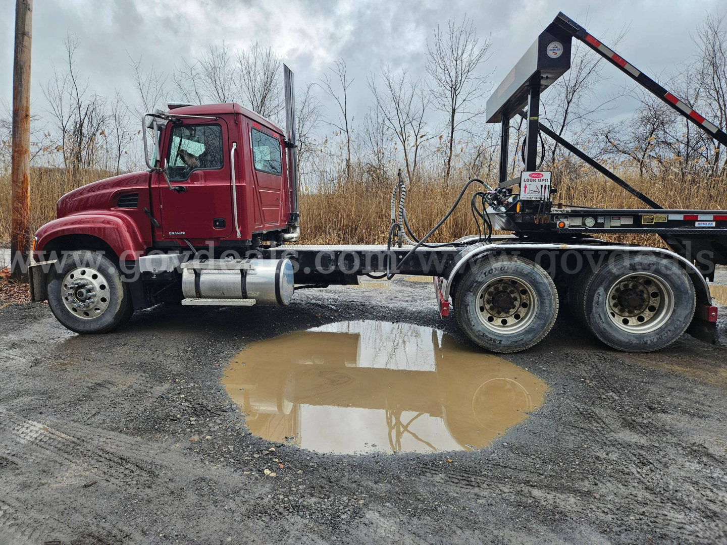 Mack CV713 Granite truck cab and rear chassis with a flatbed trailer and a wheel racked on a trailer; puddle in the foreground