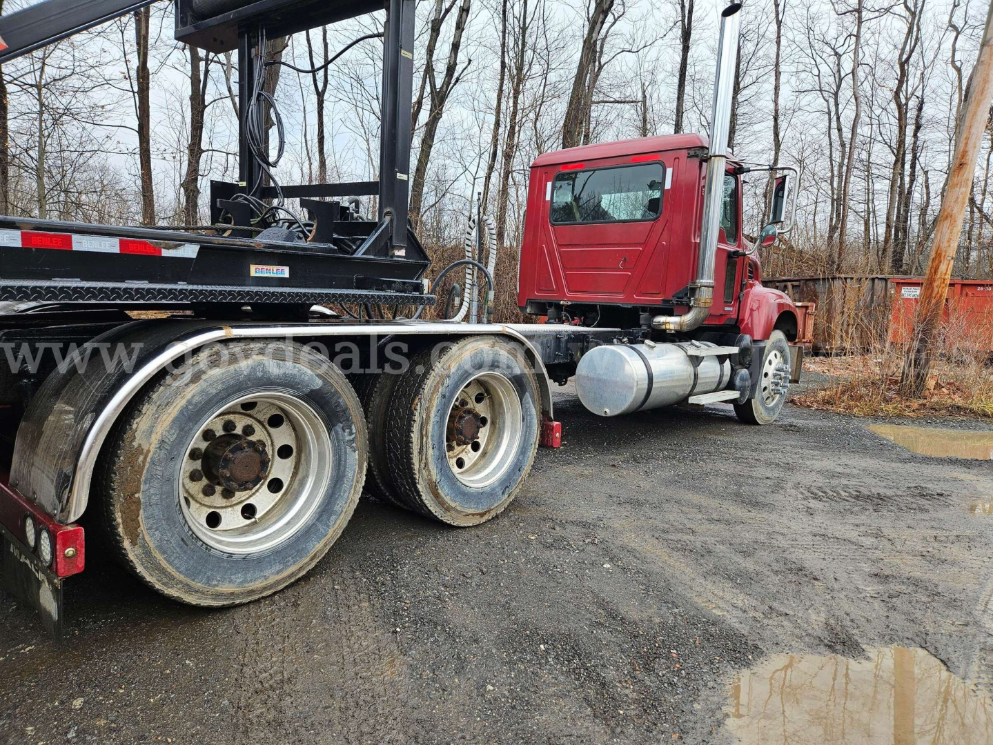 Truck tractor with red cab connected to a trailer, Mack CV713 Granite, rear tires visible, gravel lot, autumn trees in background.