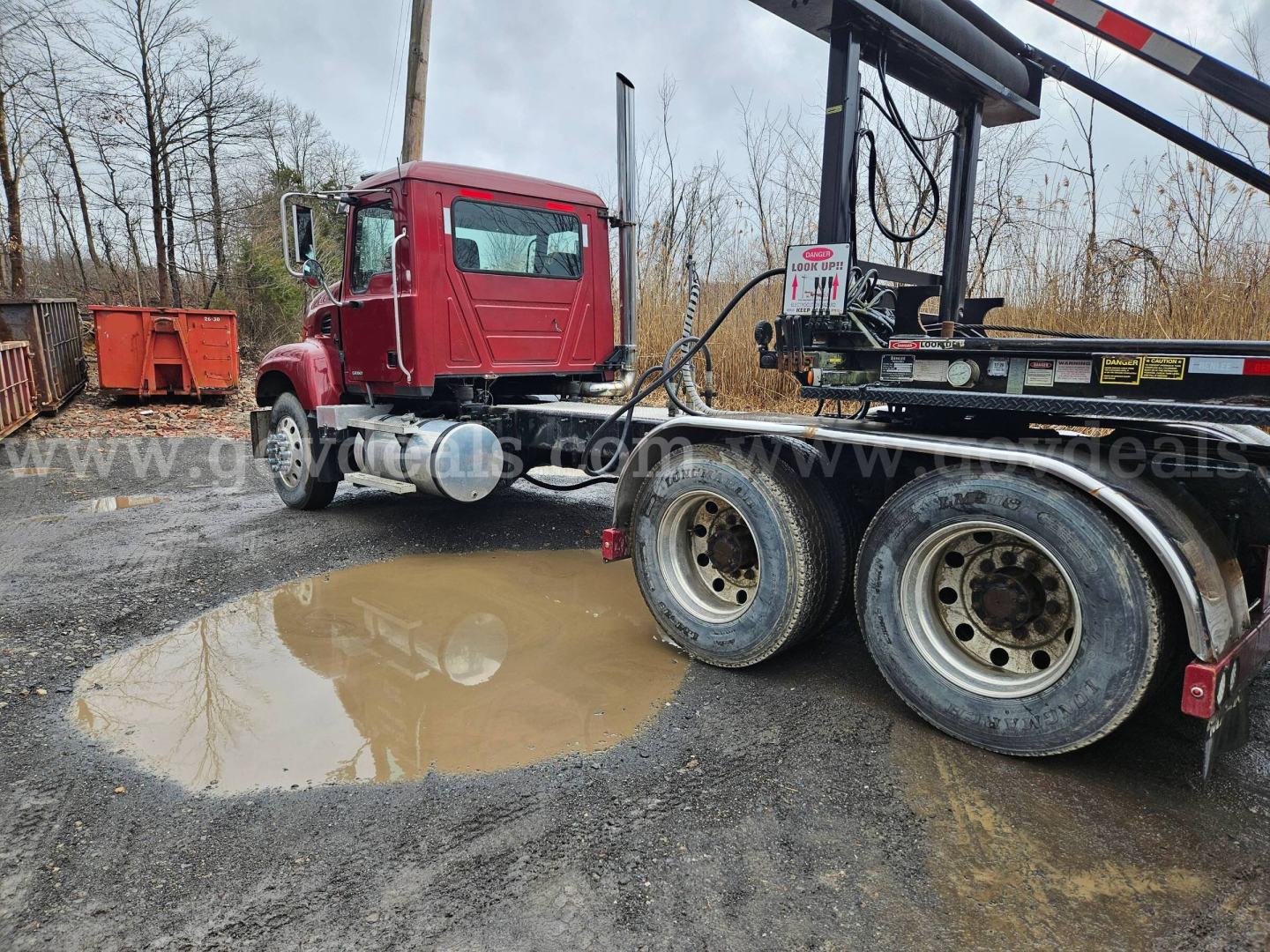 Truck cab and trailer chassis visible with a red Mack CV713 Granite truck in a salvage yard; rear wheelset and mud puddles on muddy ground.