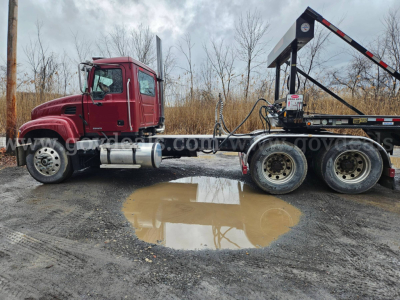 Mack CV713 Granite truck cab and rear chassis with a flatbed trailer and a wheel racked on a trailer; puddle in the foreground