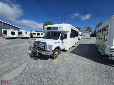 Ford E450 wheelchair bus side/front view showing 2013 and 2019 model years, bus with front bumper guard, parked on gravel lot among other buses.