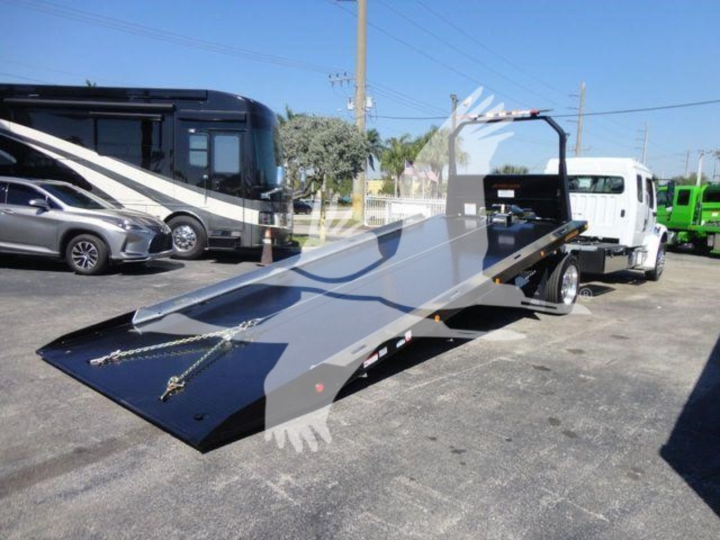 Flatbed tow truck ramp in an outdoor lot, showing a light-gray steel platform with a raised guard rail and winch pulley, side chains, and black nonslip surface.